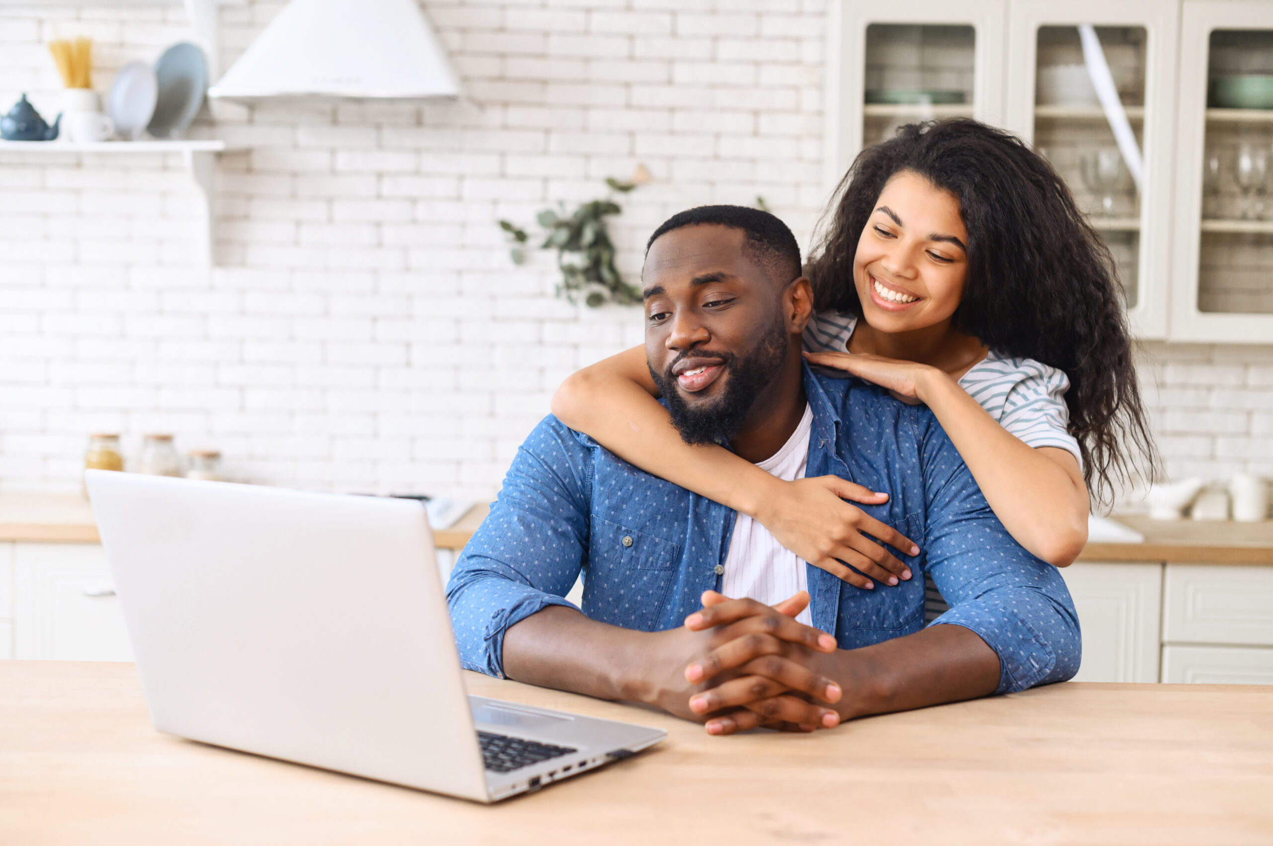 Woman hugging man from behind. The couple is relaxing in their kitchen, browsing internet on laptop about mortgage rates and finances