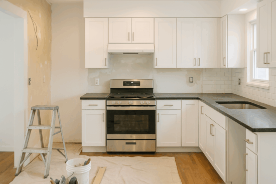 A kitchen renovation in progress featuring white shaker cabinets, black countertops, a stainless steel gas range, and an unfinished backsplash. A ladder, drop cloth, and bucket of tools are visible on the wooden floor, indicating ongoing construction work