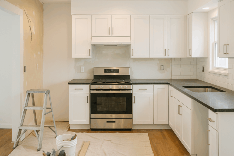 A kitchen renovation in progress featuring white shaker cabinets, black countertops, a stainless steel gas range, and an unfinished backsplash. A ladder, drop cloth, and bucket of tools are visible on the wooden floor, indicating ongoing construction work