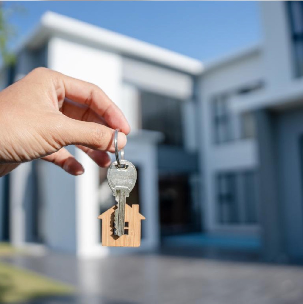 Hand holding a house key and wooden house-shaped keychain in front of a modern home.